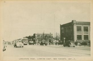 Black/white photo postcard depicting Lakeshore Road at street level with cars and a trolley car…