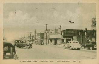 Black/white photo postcard depicting Lakeshore Road at street level with cars going by. Caption…