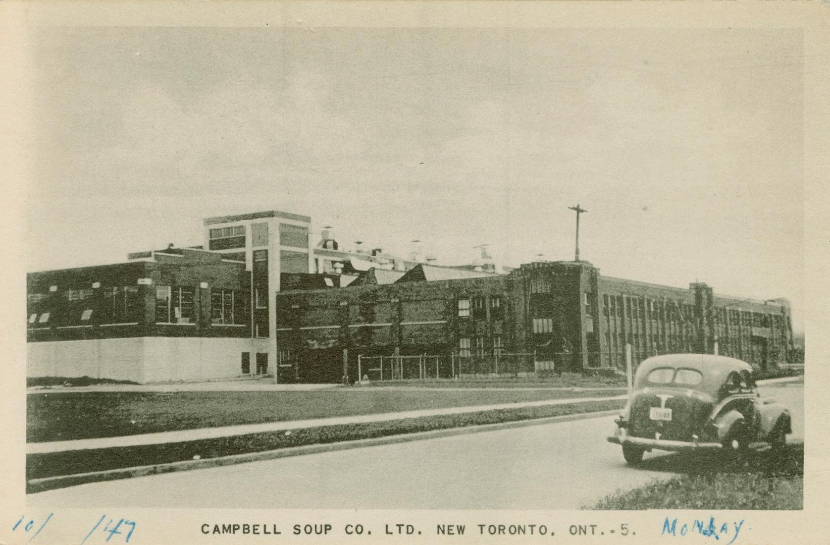 Black and white photo postcard depicting a large warehouse building with a car parked across th…