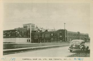 Black and white photo postcard depicting a large warehouse building with a car parked across th…