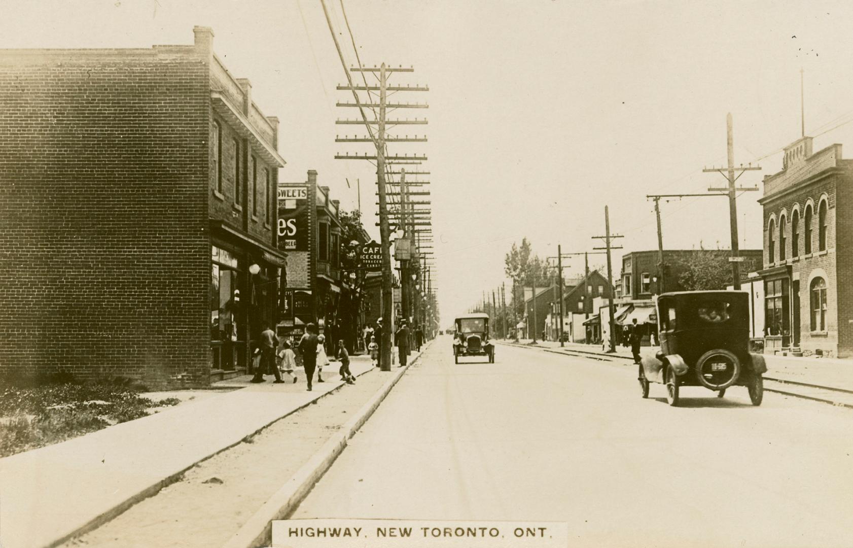 Black and white photo postcard depicting a street with cars, hydro-poles, and rail tracks in vi…