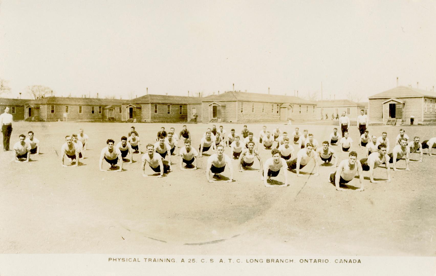 Sepia-toned photo postcard depicting a team of men doing push-ups in front of army barracks at …