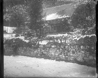 A photograph of a cottage located behind a stone fence approximately four feet tall and some tr…