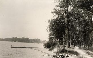 Black and white photo postcard depicting a pathway through a forest alongside a shoreline and s…