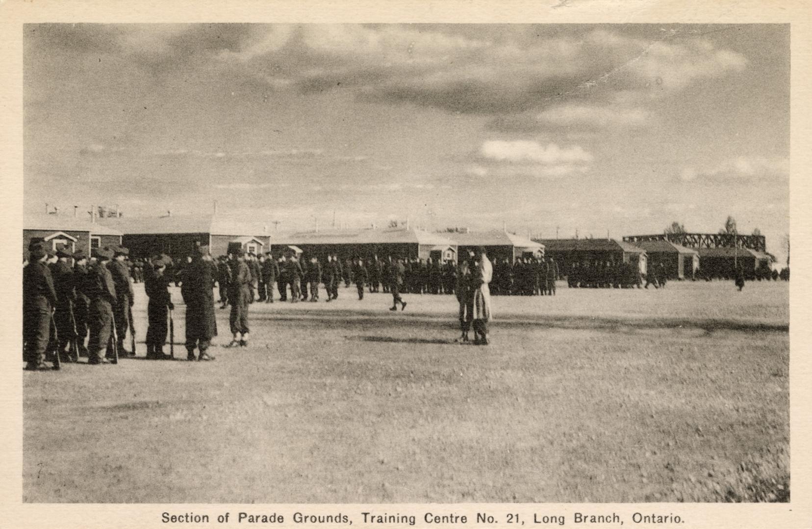 Sepia-toned photo postcard depicting men lined up on a large field in front of the barracks at …