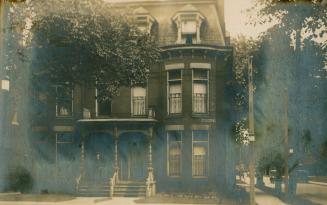 Black and white photograph a man standing in the doorway of a semi-detached three story house.