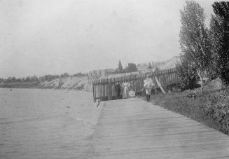 A photograph of a boardwalk beside a body of water. There are three people walking on the board…