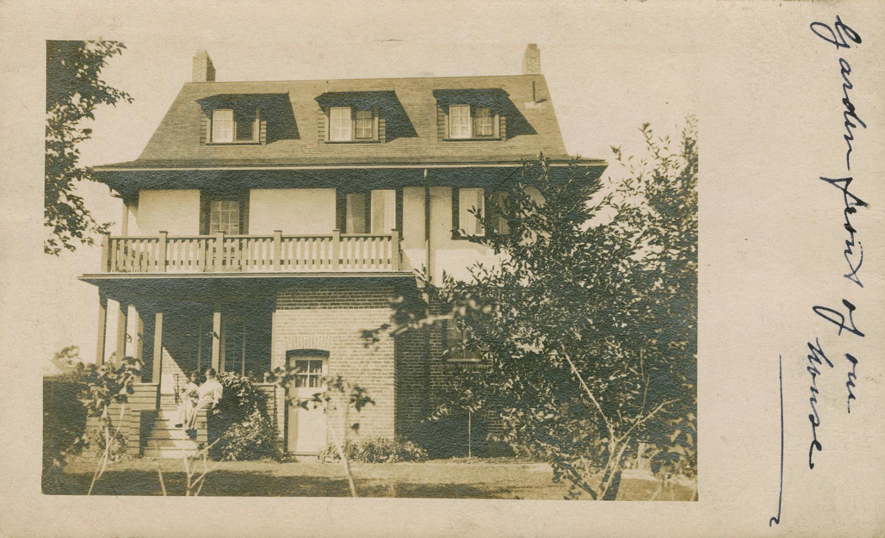 Black and white photograph of two people standing on the front veranda of a three story house.