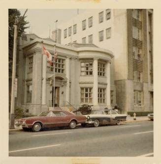 A photograph of a three story building with tall columns and large windows facing a paved city …