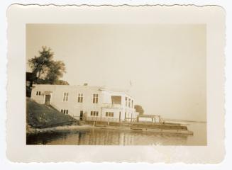 A photograph of a rowing club next to a body of water, with a dock and piers extending into the…