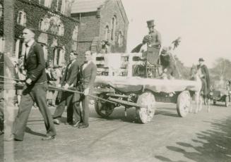 A photograph of a parade through a university campus, with people in suits and bow ties pulling…