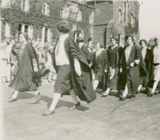 A photograph of a parade through a university campus, with people in academic robes and ties wa…