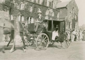 A photograph of a parade through a university campus, with a carriage being pulled by two horse…