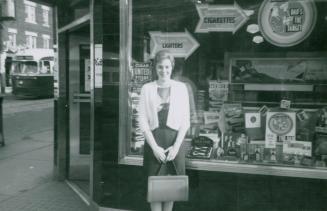 A photograph of a person standing on a city sidewalk in front of a shop window located at the c…