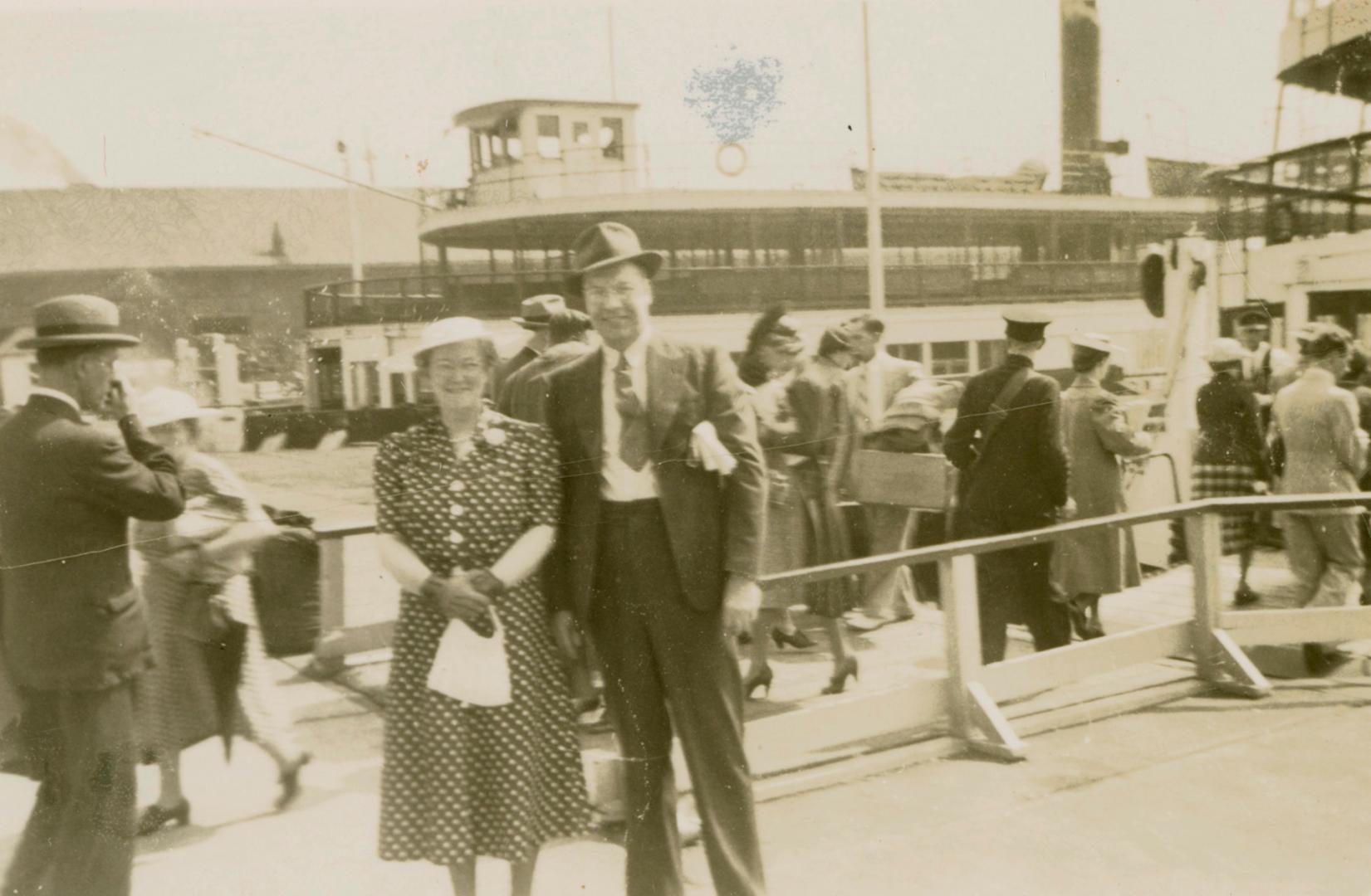 A photograph of two people posing for a photograph in front of two ferries docked in a busy har…