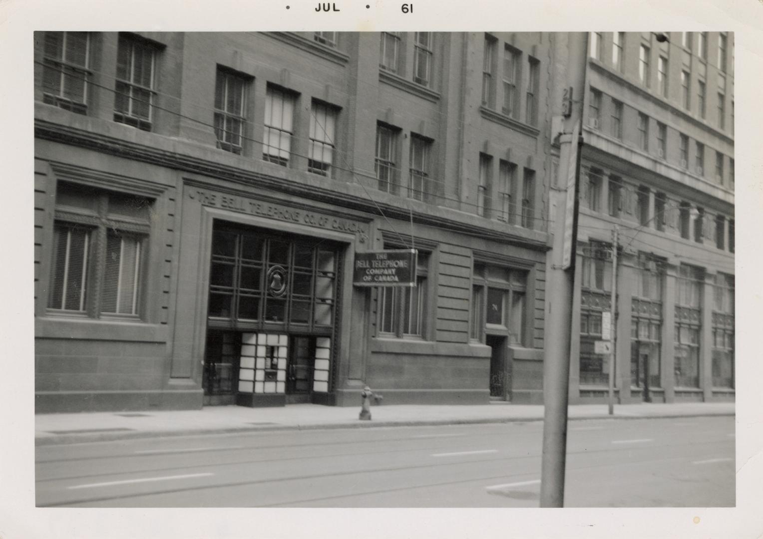 A photograph of an office building located on a city street, with a sidewalk, telephone poles a…