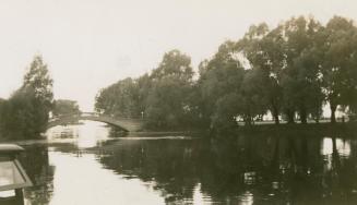 A photograph of a pedestrian bridge spanning a body of water between two islands, with trees an…