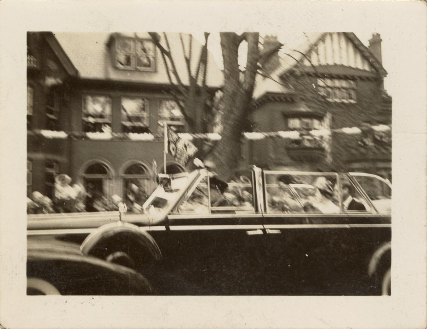 A photograph of a car driving down a city street, with a tree and houses in the background, as …