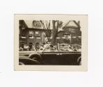 A photograph of a car driving down a city street, with a tree and houses in the background, as …