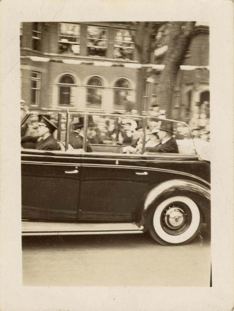 A photograph of a car driving down a city street, with large crowds of people lining the sidewa…