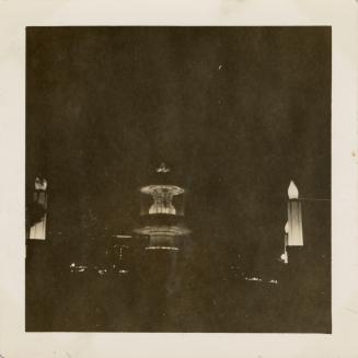 A photograph of a large round fountain with two basins, taken at night. The fountain is lit fro…