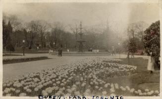 A photograph of a large public park, with flower gardens, paved pathways and a multi-tired foun…