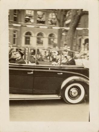 A photograph of a car driving down a city street, with a tree and houses in the background, as …