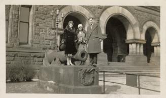 A photograph of three adults standing next to a cannon located next to arched entranceways to a…