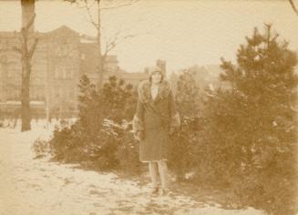 A photograph of a person standing in front of some shrugs and short pine trees. There is a larg…