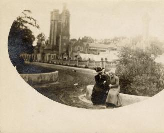 A photograph of two people sitting on a ledge next to a curving path or road leading towards a …