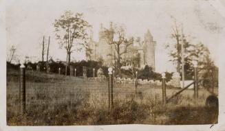 A photograph of a stone castle-like building, with a wire fence, trees and grassy hill in the f…