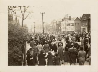 A photograph of a large crowd of people walking on and next to a paved city street, with houses…