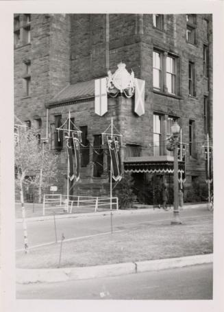 A photograph of a large brick building, with banners hung on posts in front of it and crests at…