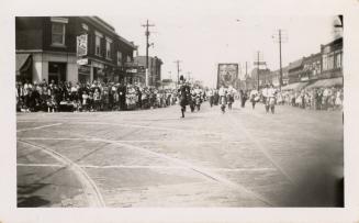 A photograph of a parade in the middle of a paved city street, with many spectators lining the …