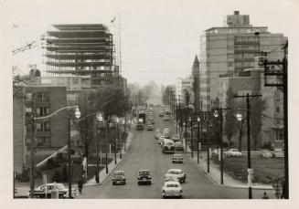 A photograph of a wide, paved city street taken from a slightly elevated vantage point. There a…