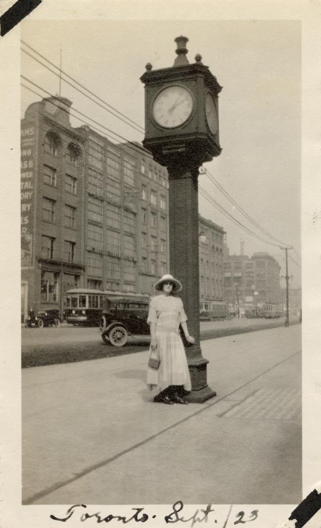 A photograph of a person posing in front of a clock on a tower approximately twenty feet tall, …