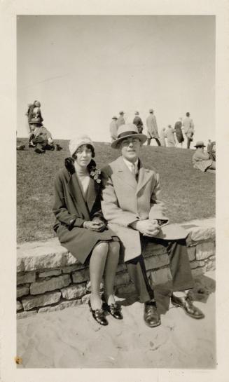 A photograph of two people sitting on a stone ledge in front of a small grass hill. There are o…