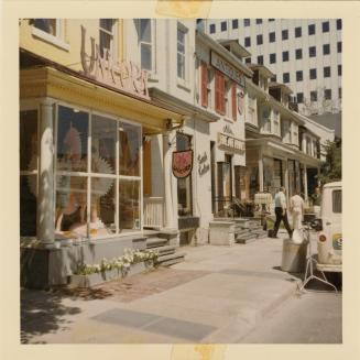 A photograph of a city street, with two and three story buildings on the left side of the road  ...