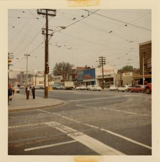 A photograph of a city street and intersection. There are crosswalks painted at two corners of …