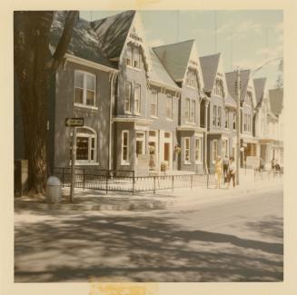 A photograph of a paved city street, with a row of houses on the right side of the street. Ther…