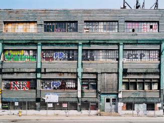 A photograph of a three story warehouse behind a chain link fence, with a sign on the fence ind…