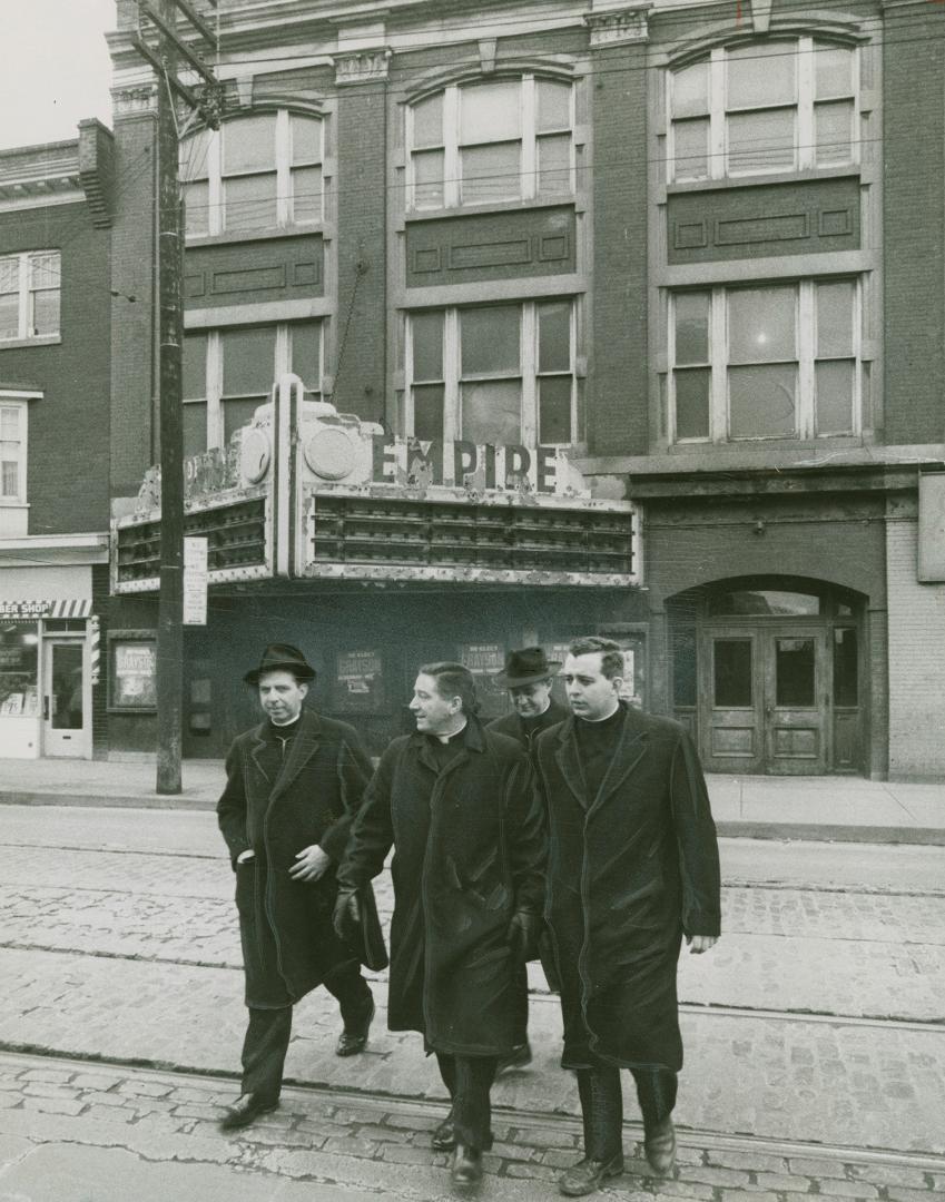 A photograph of four men walking across a paved city street, which has bricks exposed and stree ...