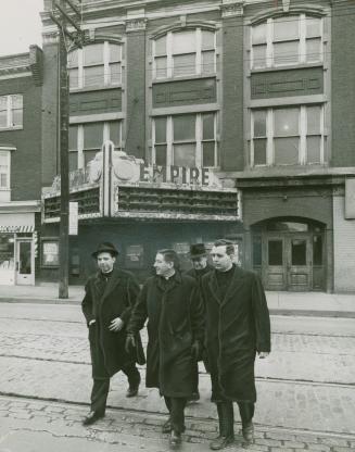 A photograph of four men walking across a paved city street, which has bricks exposed and stree…