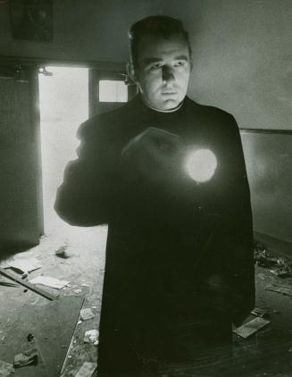 A man in a black robe shines a flashlight to the right of the camera. Through a door, the sun s…