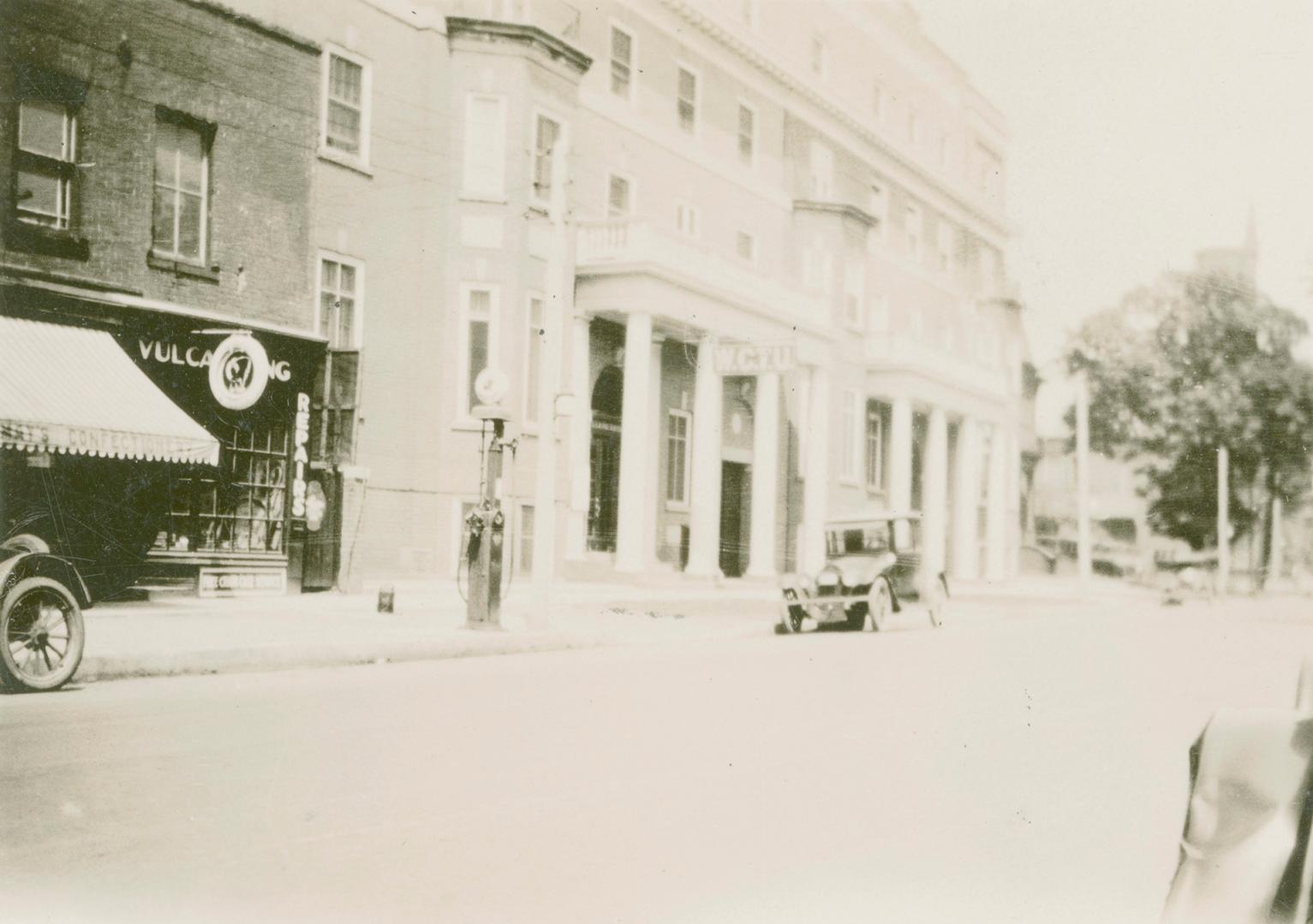 A photograph of a city street, with a paved road and a row of two, three and four story buildin ...