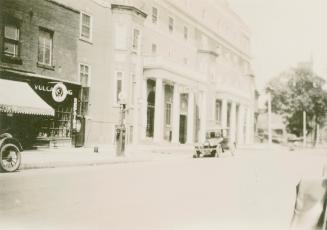 A photograph of a city street, with a paved road and a row of two, three and four story buildin…