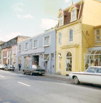 A photograph of a paved city street with a row of two and three story buildings on the far side…