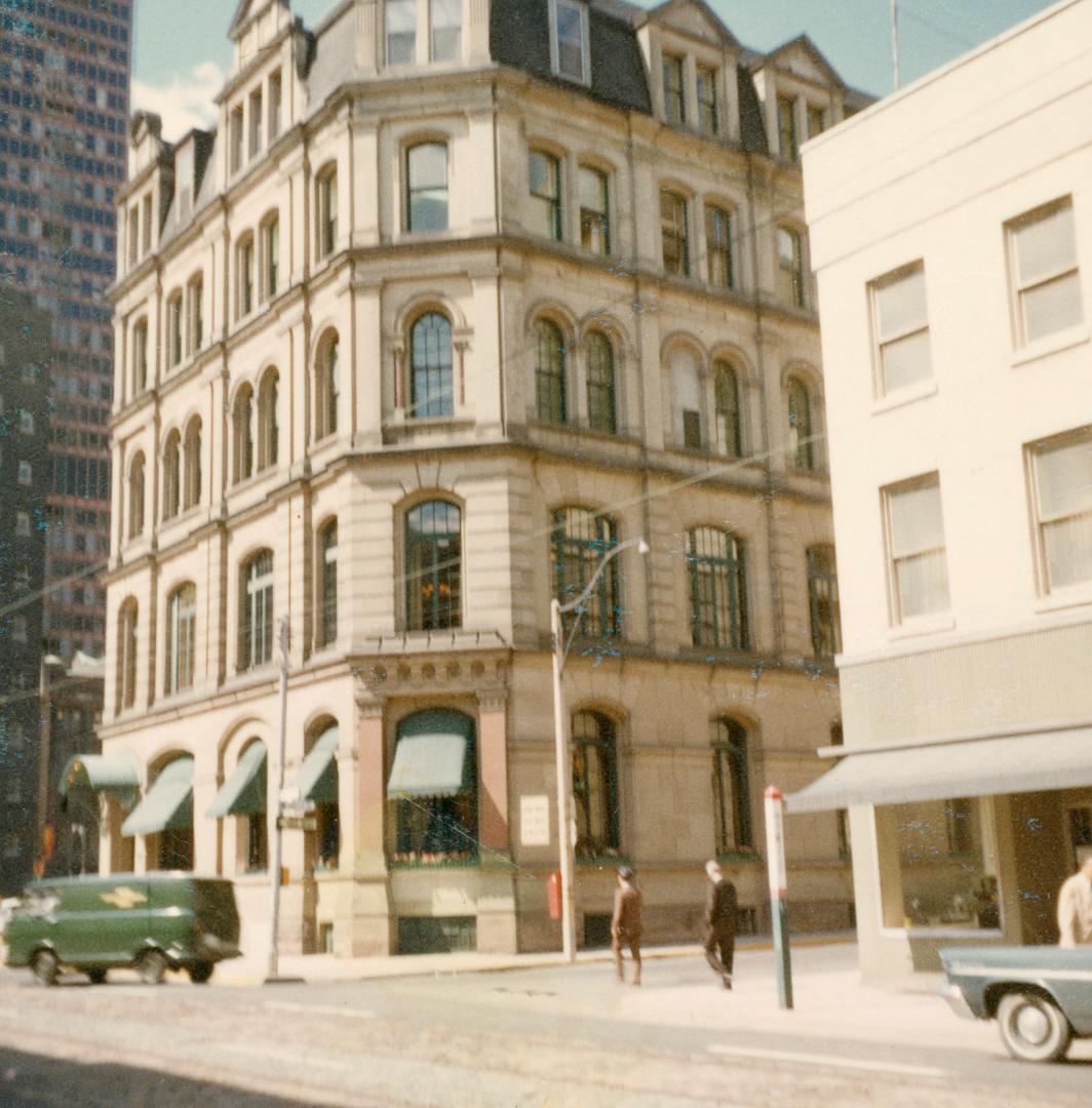 A photograph of a five story building at the corner of a city intersection. There is a paved ro…