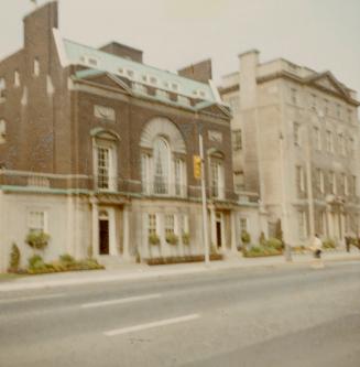 A photograph of two three or four story brick or stone buildings beside a paved city street. Th…