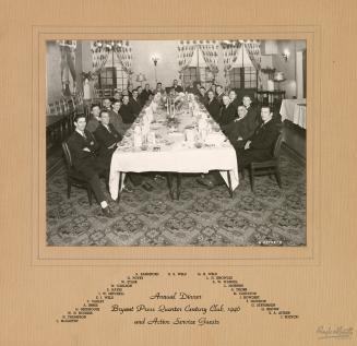 A photograph of a group of people posing around a long banquet table, which is set with dinnerw…